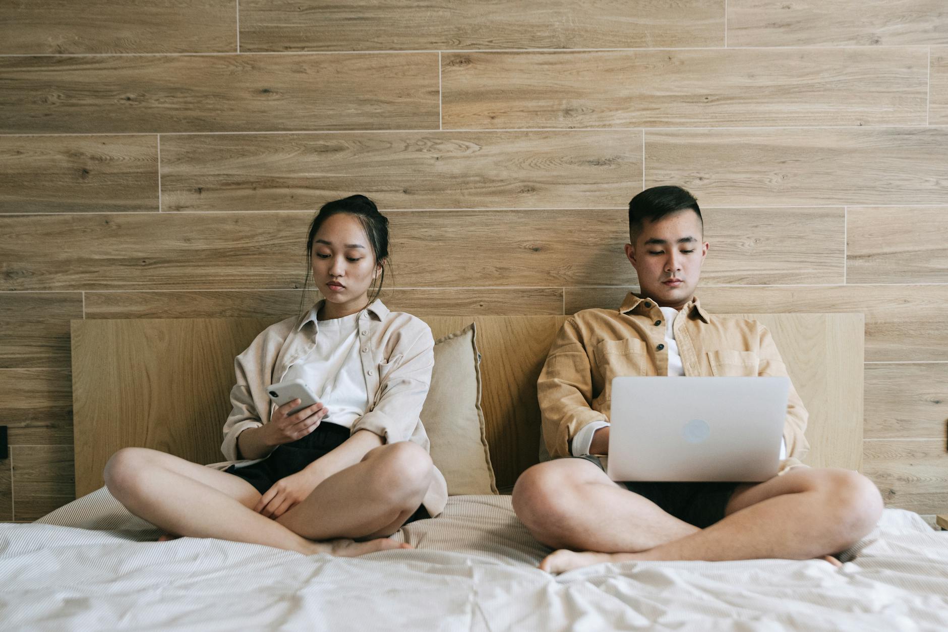 man and woman sitting on the bed while using gadgets